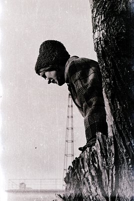 Black-and-white portrait of a person in winter attire—knit cap, scarf, and checkered coat—leaning against a rough tree trunk....