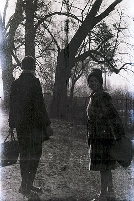 Black-and-white street scene featuring two figures in early 20th-century attire—man in a long coat and hat, woman in a long c...