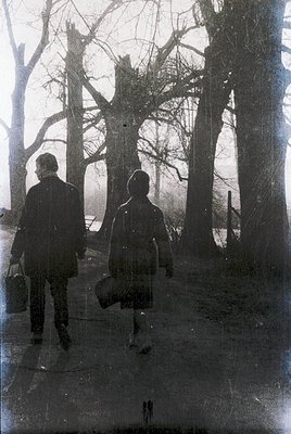 Two figures in vintage winter attire walk a snow-covered path through a leafless forest, silhouetted against bare trees. The ...