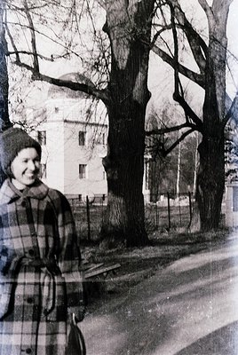 Vintage black-and-white portrait of a woman in a plaid coat and knit hat, standing beside a tree-lined street. Mid-20th centu...