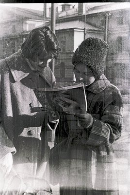 Vintage black-and-white photo of two individuals sharing a book in an urban setting, likely mid-20th century. The adult wears...
