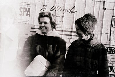 Two women in winter attire pose against a backdrop of vintage European spa posters, likely from the 1960s–1970s. The woman on...