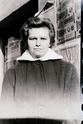 Portrait of a woman in a mid-century winter coat with a fur collar, standing indoors near a wall adorned with printed fabric ...
