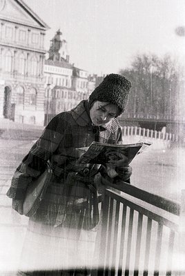 A person in a vintage plaid coat and knit cap reads an open book while seated on a wrought-iron bridge railing, set against a...