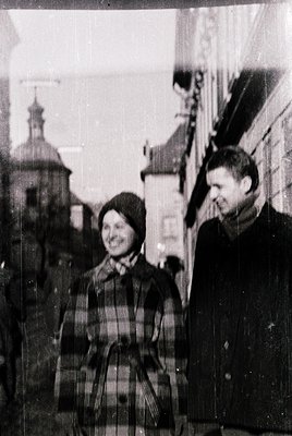 Vintage black-and-white street portrait of two individuals in winter attire: woman in plaid skirt, knitted hat, and scarf; ma...