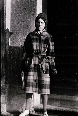 Young woman in a 1950s-60s plaid coat and knit hat poses outdoors near a building entrance. The checkered pattern and high-co...