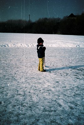 Child in winter attire stands alone on snow-covered field under starry night sky. Dark coat, yellow pants, and hat suggest mi...