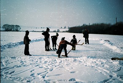 Vintage winter scene of a makeshift ice rink in a rural field, featuring six people—likely mid-20th century—engaged in inform...