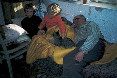 A candid 1970s-era indoor scene captures three generations sharing a moment on a patterned rug. A man in a striped shirt and ...