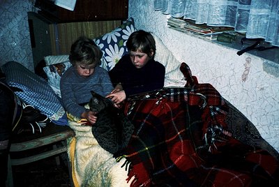 Two children play with a cat on a bed covered in patterned blankets, likely mid-20th century. The boy holds the cat while the...