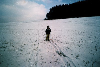 A lone cross-country skier pauses mid-stride on a groomed trail, dressed in vintage winter gear (beanie, goggles, padded jack...