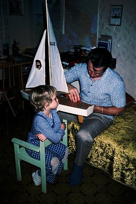 Indoor scene featuring a man and child assembling a model sailboat (DJX logo) in a mid-20th-century home. The man, seated on ...