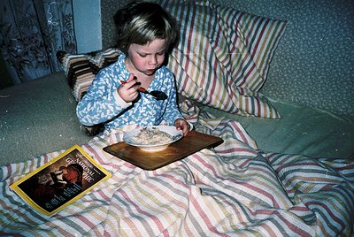 Young child eating cereal from a wooden tray on a striped bedspread, mid-1970s. Patterned pajamas with floral motifs and a vi...