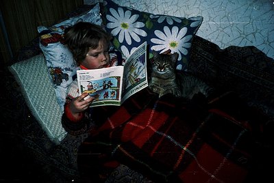 Child engrossed in a vintage comic book, seated on a patterned sofa with floral pillow. Gray tabby cat perched beside them, o...