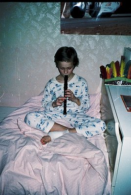 Child in vintage floral pajamas plays a recorder on a bed with patterned wallpaper, 1960s-1970s. Side table holds colorful pl...