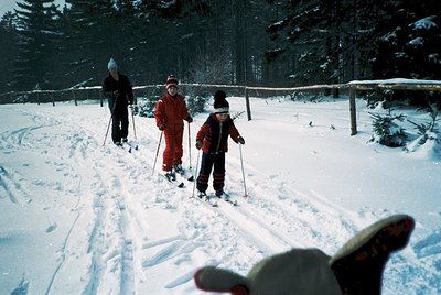 Three cross-country skiers glide through snow-covered forest, dressed in 1970s-style winter gear. Bright orange and red suits...