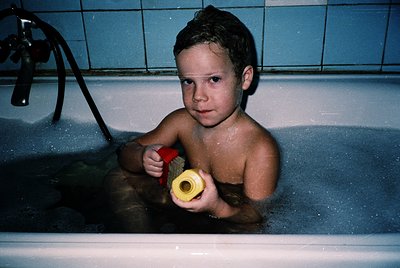 Young boy in a vintage bathtub holding a yellow plastic toy, likely a 1970s–1980s household scene. Blue-tiled walls and chrom...