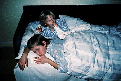 Two children rest on a twin bed in a dimly lit room, likely mid-20th century. The girl on top holds a stuffed animal, while t...