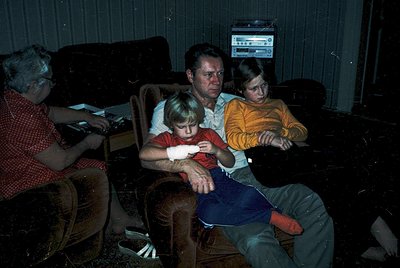 Vintage family portrait in a dimly lit living room, featuring four individuals seated on upholstered furniture. A man and wom...