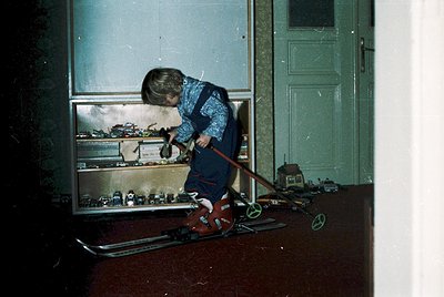 Child in vintage indoor setting, organizing toy train set on wooden floor. Display case showcases miniature vehicles, buildin...