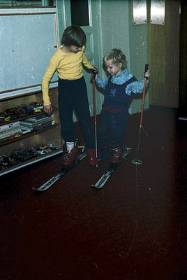 Two children practice skiing indoors, holding wooden poles and wearing vintage ski boots with "NORDICA" branding. The boy in ...