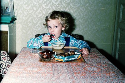 Vintage indoor shot of a young child eating a chocolate-covered doughnut at a patterned tablecloth. Mid-20th century kitchen ...