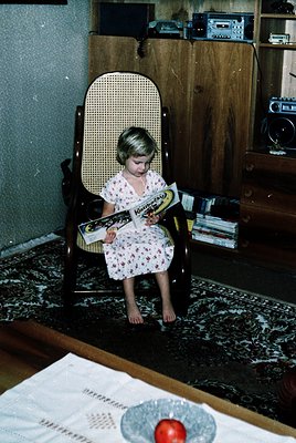Vintage mid-century interior featuring a young girl in a floral dress reading a comic book titled *"Kondor"* in a cane-backed...