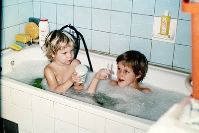 Two children play in a white-tiled 1970s-style bathroom bathtub, surrounded by vintage products: a yellow bottle (likely sham...