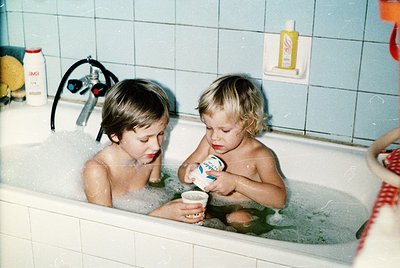 Two young boys play in a white-tiled bathtub, splashing water. One holds a small cup, the other reaches toward it. Mid-20th-c...