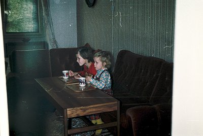Two children share a moment over tea in a mid-20th-century interior. The girl in a floral dress holds a cup, while the younge...