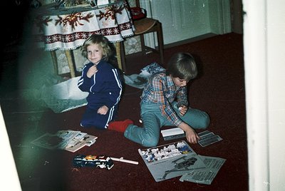 Two children assemble model kits on a carpeted floor, surrounded by scattered parts. Girl in a 1980s-style tracksuit poses be...
