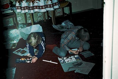 Two individuals assemble model kits on a patterned carpet in a dimly lit room, likely mid-1970s. Decorative embroidered curta...