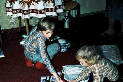 Vintage 1970s indoor scene: two children playing with wrapped gifts on a patterned carpet. Boy in plaid shirt/knee-length pan...
