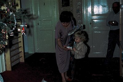 Vintage indoor holiday scene featuring a woman in a long dress holding a child in a checkered shirt, both examining a wrapped...