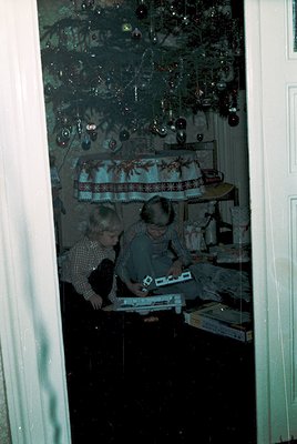 Two children play with a wooden train set on a table draped with a vintage floral tablecloth, surrounded by holiday decoratio...