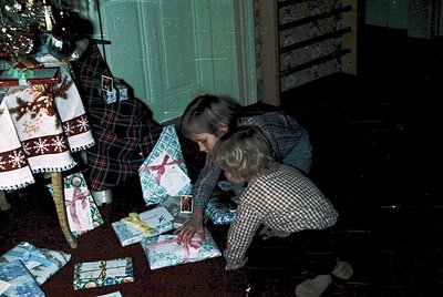 Two children kneeling on a carpeted floor, opening wrapped holiday gifts in a cozy indoor setting. Patterned wallpaper and a ...