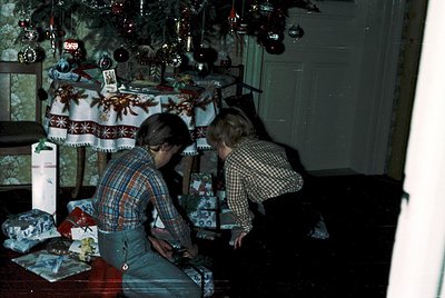 Two children decorate wrapped gifts under a festively adorned Christmas tree, 1970s–1980s. Patterned tablecloth, snowflake or...