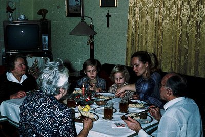 Family dinner scene in a mid-century interior, featuring six adults and children seated around a table. A vintage TV, wall-mo...
