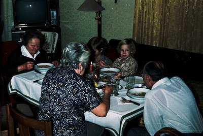 Family meal in a mid-century dining room, featuring five adults seated around a round table with white tablecloths. Visible a...