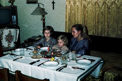 Family gathers around a set table in a 1970s-style home, adorned with vintage tableware and a small Christmas tree. Mother an...