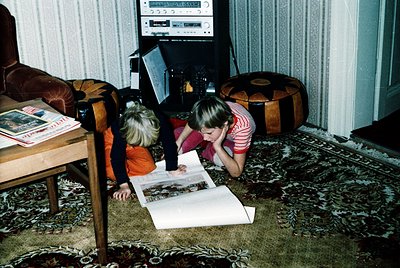 Two children engrossed in a large open book on a vintage patterned carpet, surrounded by mid-century furniture. The boy in or...