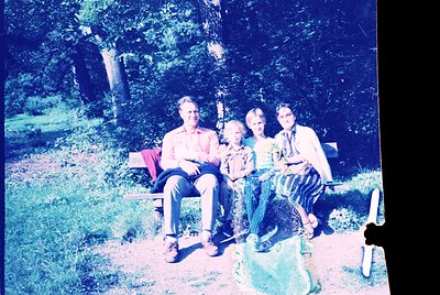 Vintage sepia-toned family portrait in a wooded park setting, mid-20th century. Four individuals seated on a wooden bench, su...