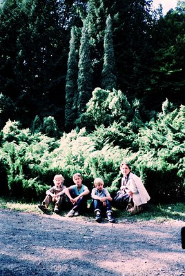 Four children and an adult pose outdoors on a gravel path surrounded by manicured evergreen shrubs. The adult wears a light c...