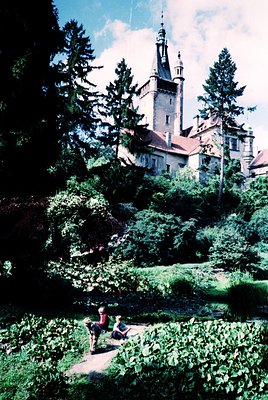 Historic castle perched on a hillside, surrounded by lush greenery and mature coniferous trees. Two figures sit on a stone br...