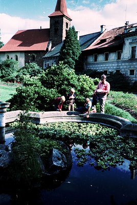 Family of four—adult and three children—standing beside a manicured hedge maze in a courtyard. Red-tiled roof and stone churc...