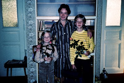 Family portrait in a dimly lit interior, likely 1970s–1980s. Three generations pose in a hallway with floral-patterned wallpa...