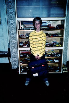 Young boy in striped sweater poses beside a glass-fronted toy display shelf filled with vintage cars, 1970s-80s. Indoor setti...
