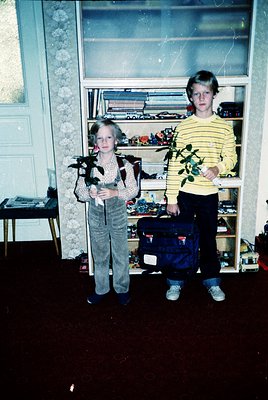 Two children pose indoors, likely mid-20th century (1960s–1970s). Boy wears striped sweater, girl in patterned sweater with b...