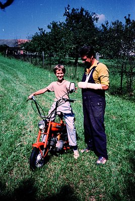 A man in a yellow shirt and dark overalls assists a smiling boy in beige shorts and a white shirt on an orange moped in a lus...