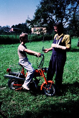 Mid-20th century rural scene: A young boy in a striped shirt rides a red lawnmower, guided by an adult in overalls and a cap....